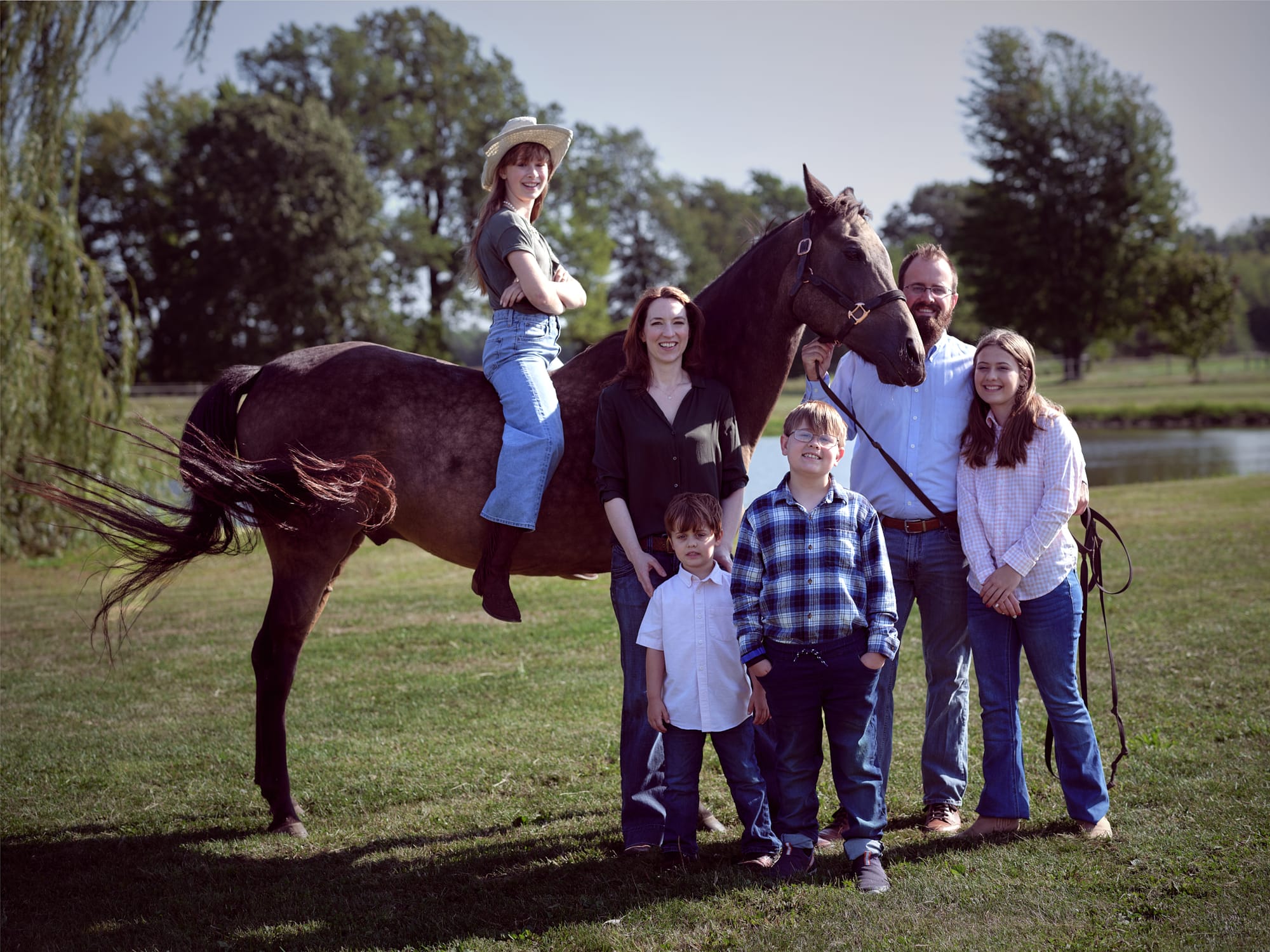 Family in front of a pond with a horse. The family is grouped to the right and one family member is sitting on top of the horse